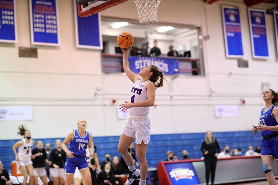 A basketball player wearing an N.Y.U. team uniform jumps up from the ground, holding a basketball. Her right hand is reaching for the hoop. Other players run toward her.