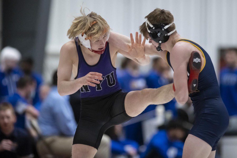 A+blonde-haired+male+wears+wrestling+protection+gear+on+the+head+and+a+purple+leotard+that+reads+%E2%80%9CN.Y.U%E2%80%9D+in+black+letters%2C+while+kicking+a+brown-haired+male+wrestler+in+a+dark+blue+leotard+with+his+left+foot.
