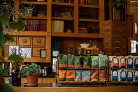 Stacks of packaged coffee beans on a wooden counter.  Behind the counter are wooden cabinets in which documents are kept.  Next to the coffee beans is a pot with plants with green leaves.