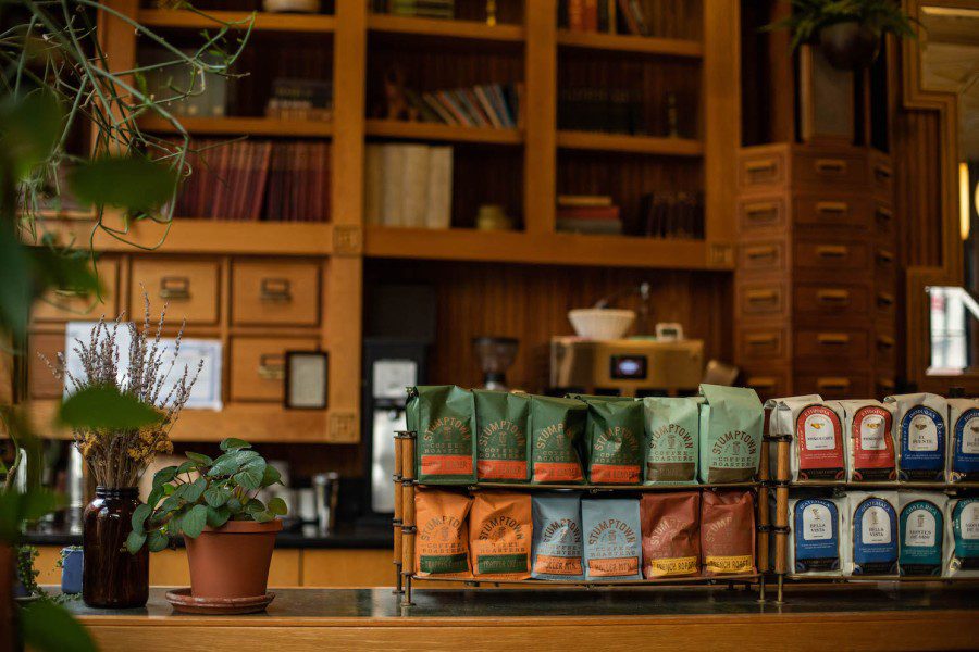 On+the+wooden+counter+are+stacks+of+pre-packaged+coffee+beans.+Behind+the+counter+are+wooden+cabinets+storing+documents+inside.+Next+to+the+coffee+beans+is+a+pot+of+plants+with+green+leaves.
