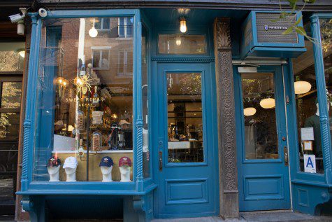 The exterior of Sweet Rehab. Blue wooden frames surround window panes. On the window pane to the left is a symbol of a bee printed in gold and texts that read “SWEET REHAB BAKERY,” also in gold.