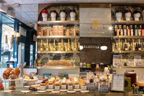 The interior of Sweet Rehab. A marble stone counter top is filled with plates of cakes, loafs of bread and cookies. On the shelves on the wall are champagne bottles and hats of various colors sitting on top of white mannequin heads. In the middle is a mirror with a gold decal that reads “SWEET REHAB NEW YORK” on it.