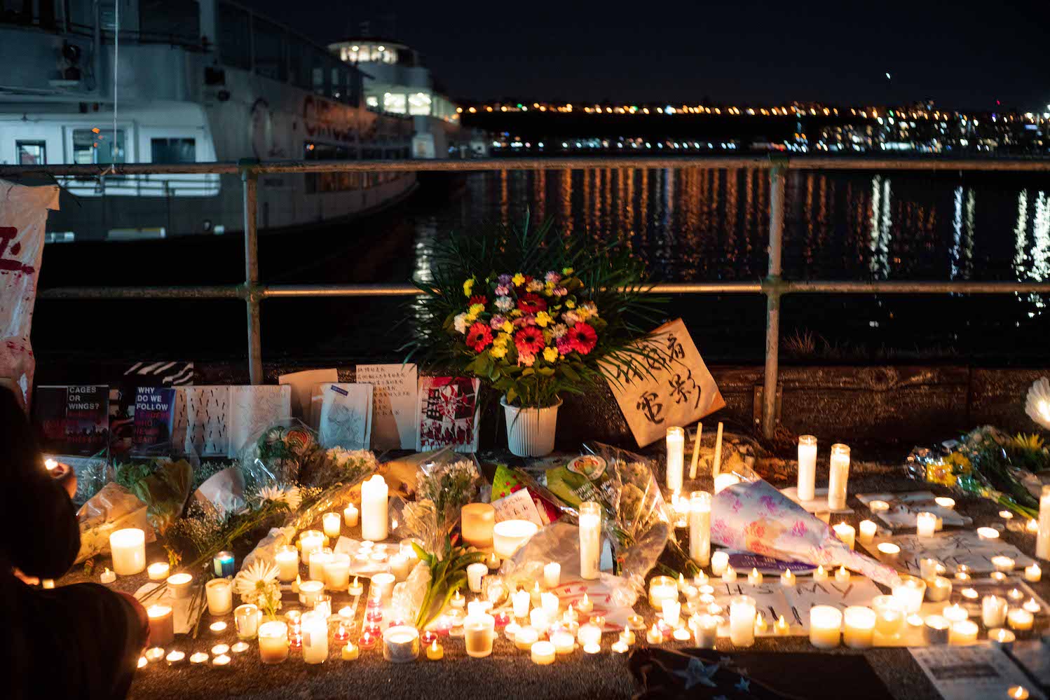 An arrangement of flowers and candles laid on the ground with commemorative texts in Chinese written on posters and papers around the candles.