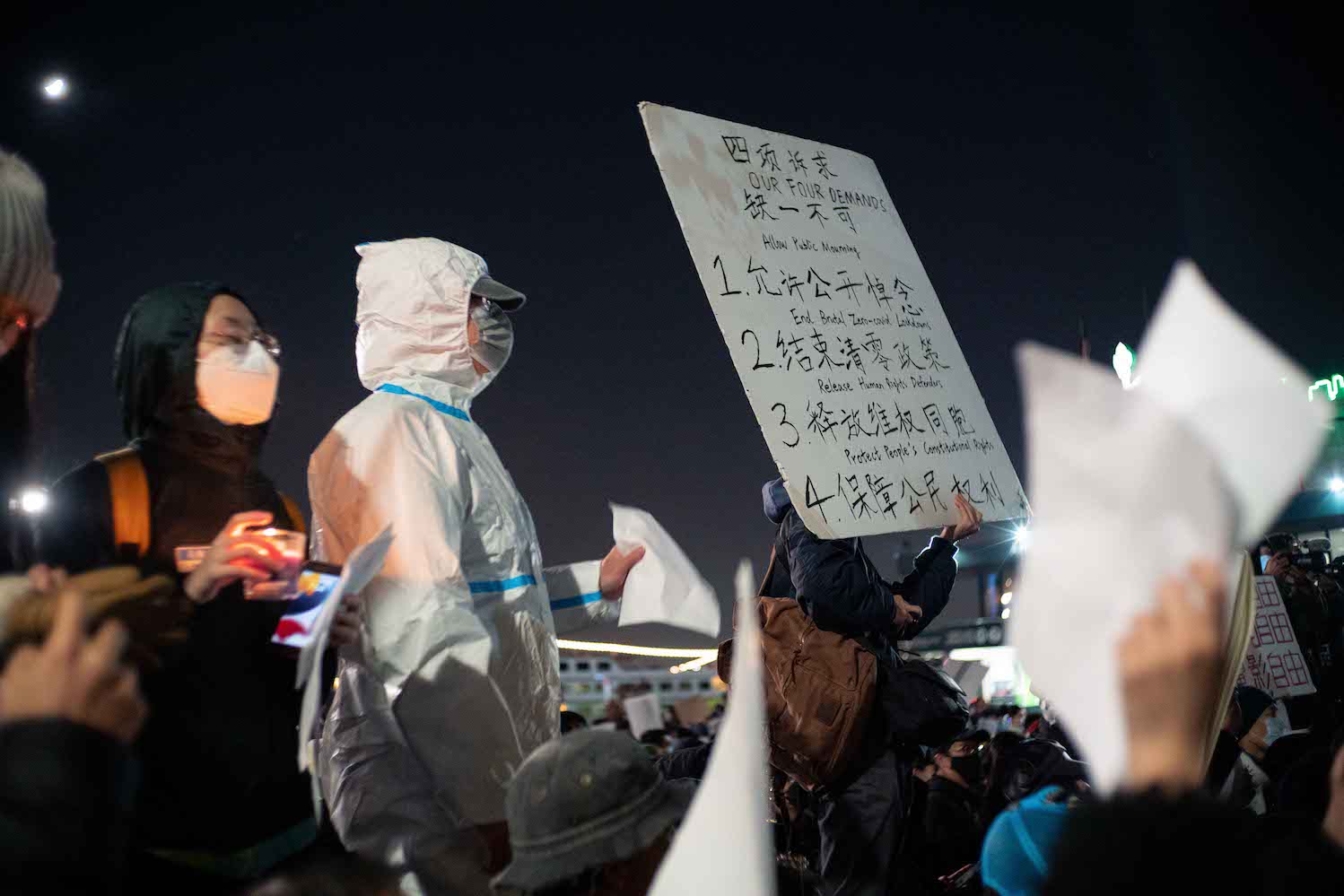A group of protesters hold signs at night that translate to “Our Four Demands, not one can be missed.” A protester is dressed in a white medical suit used by medical professionals during P.C.R. tests.