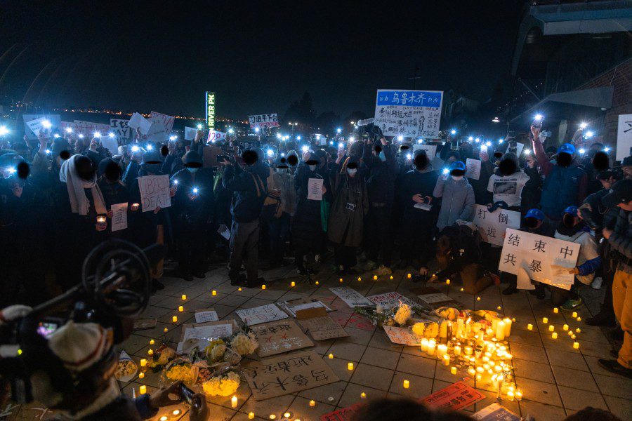 A group of protesters, mostly masked — raise their phones with the flashlights on — surrounding an arrangement of candles and flowers.