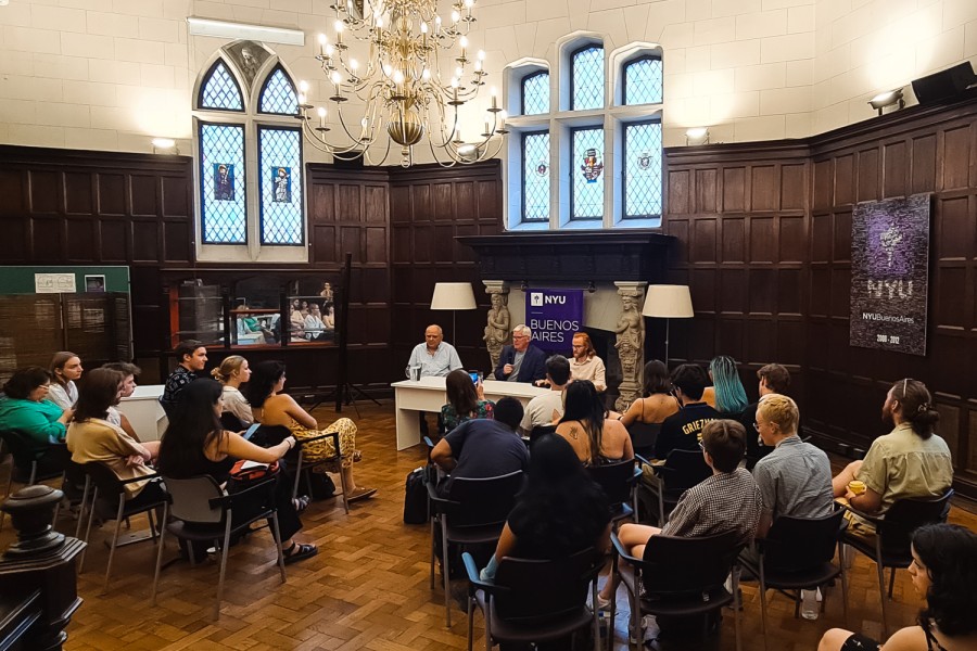 A group of people sits in a room with a chandelier and dark wood padding on the wall. In the middle of the room are three people sitting behind a white desk talking to the audience. Behind them is a poster with text N.Y.U and Buenos Aires.