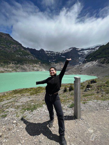 Juliana Guaraccino wears a black sweater and black jeans. She poses in front of a lake surrounded by mountains.