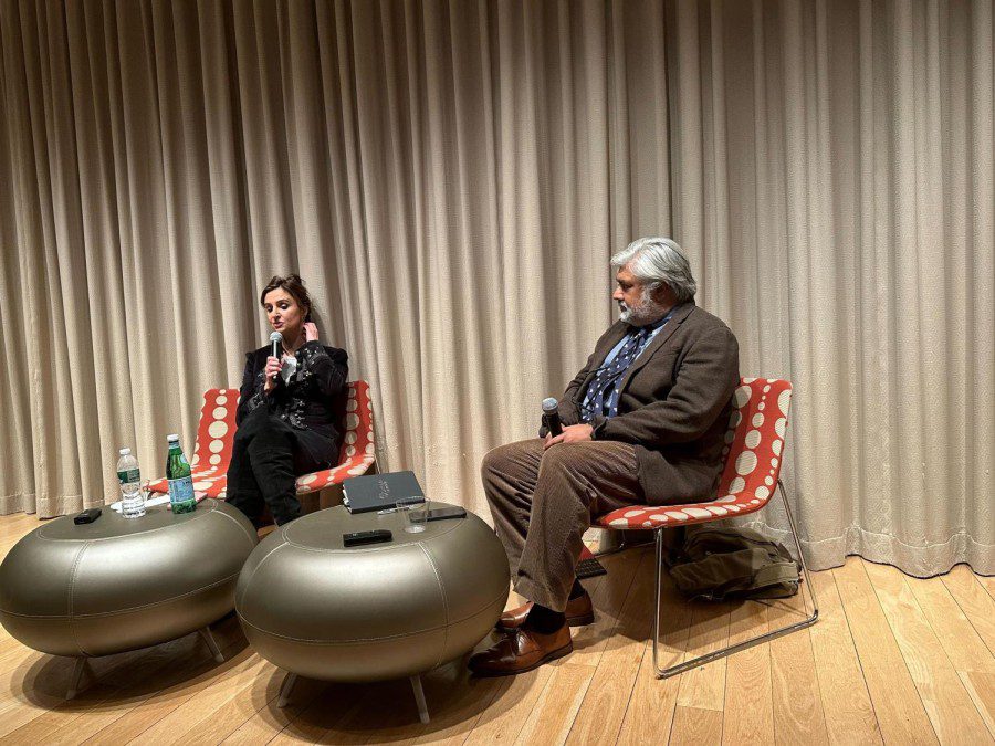 A woman wearing all black sits in a red chair with khaki polka dots as she holds a microphone. To her right, a man wearing a brown suit, a blue shirt, and a blue tie also sits in a red chair with khaki polka dots as he holds a microphone. In front of them, there are two small light brown round tables.