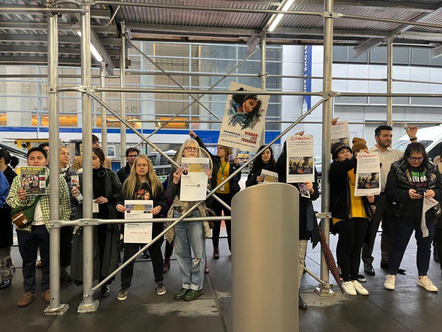 An image of multiple people standing under gray scaffolding. They are wearing yellow, black, and white shirts, and they hold white signs. Behind them, there is a gray building for The New York Times.
