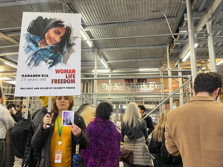 Multiple people stand under gray scaffolding. In the background, there is an entrance to a building with a sign that reads “The New York Times. A woman wearing a yellow shirt holds a sign that displays an image of a female with blood on her face. Under this image it reads “Hananeh Kia,” “23 years old,” and next to this, it reads in red font “Woman,” “Life,” “Freedom.” At the bottom of the poster in black font it reads: “Was shot and killed by security forces.”
