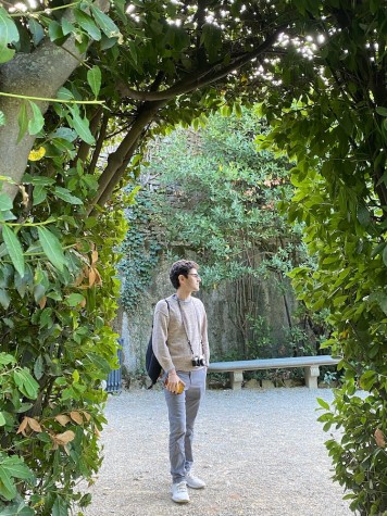 Luca Richman wears a light beige sweater, gray pants and a black backpack. He straps a film camera around his neck. He stands in a garden.