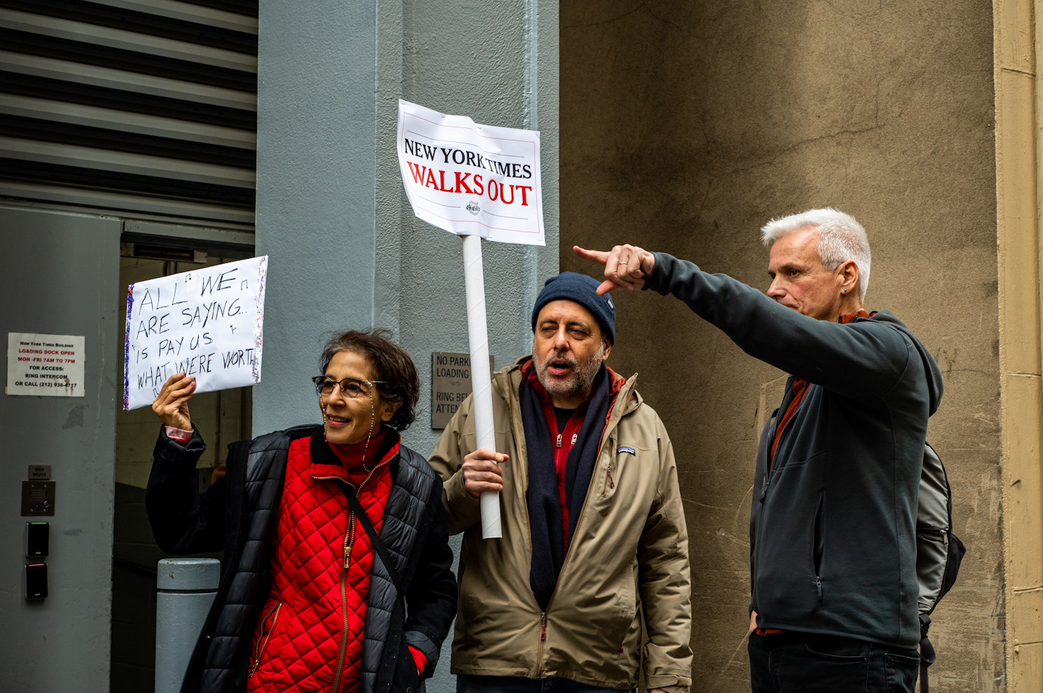 Two men and a woman standing outside of The New York Times building. A woman holds a sign that reads “All we are saying is pay us what we’re worth.” One man holds a sign that reads “New York Times Walks Out.” One man is pointing to the crowd.