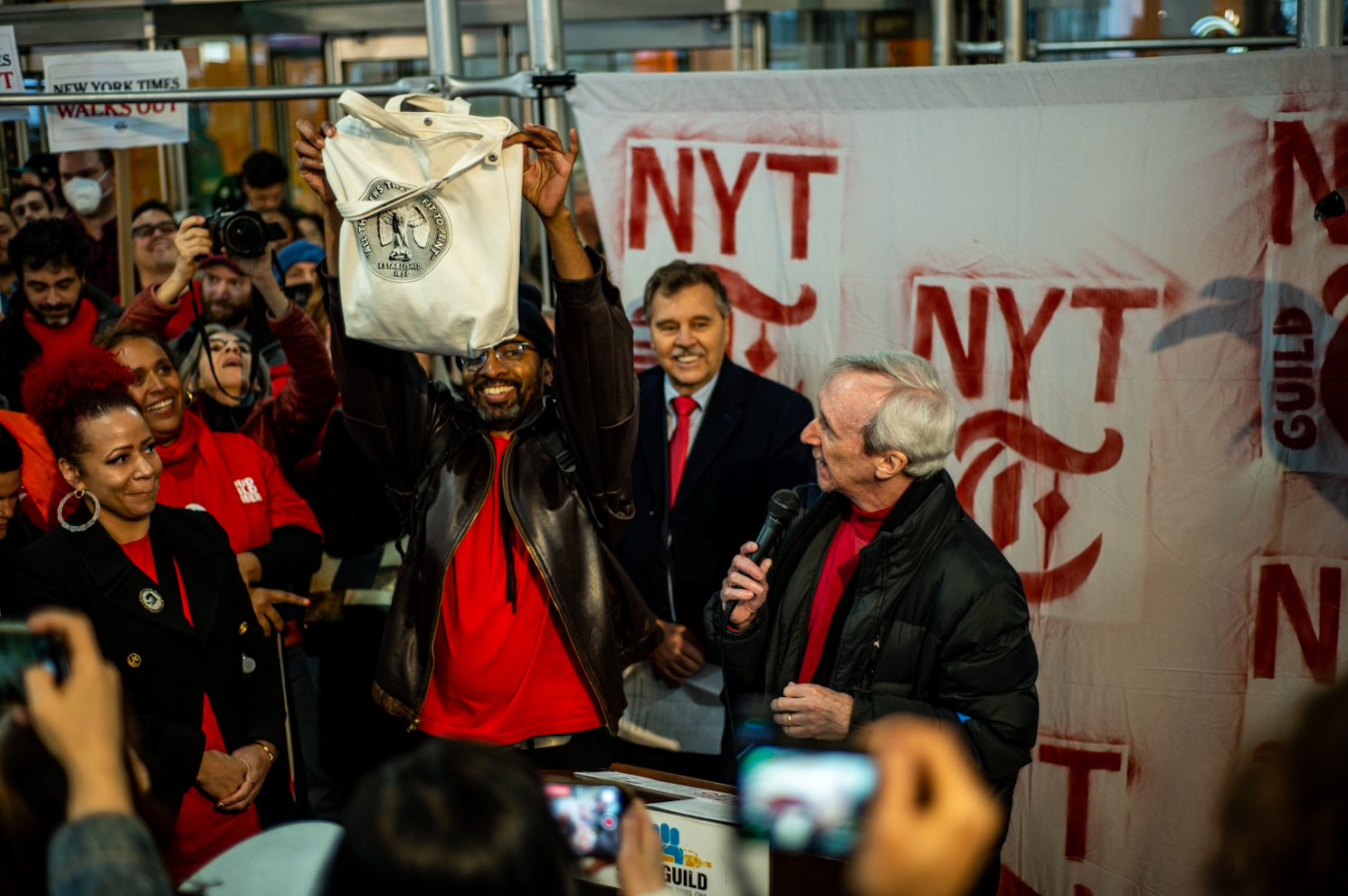 A speaker standing at a podium and a man next to him holding a New York Times tote bag. The New York Times logo and a crowd are behind them.