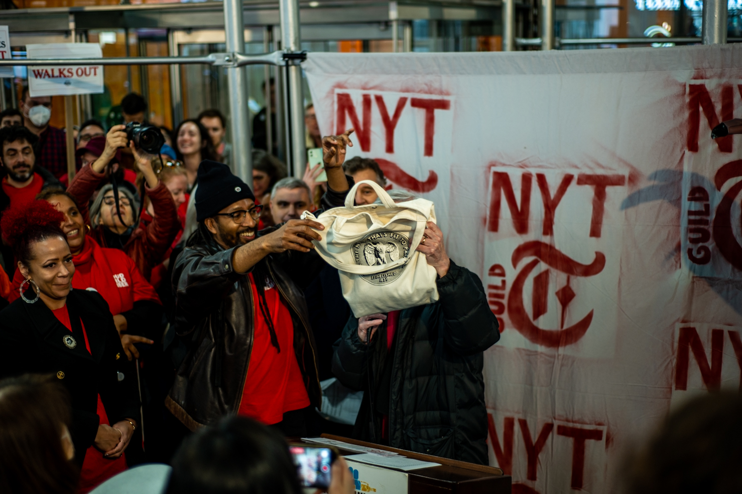 A speaker standing at a podium and a man next to him holding a New York Times tote bag. The New York Times logo and a crowd are behind them.
