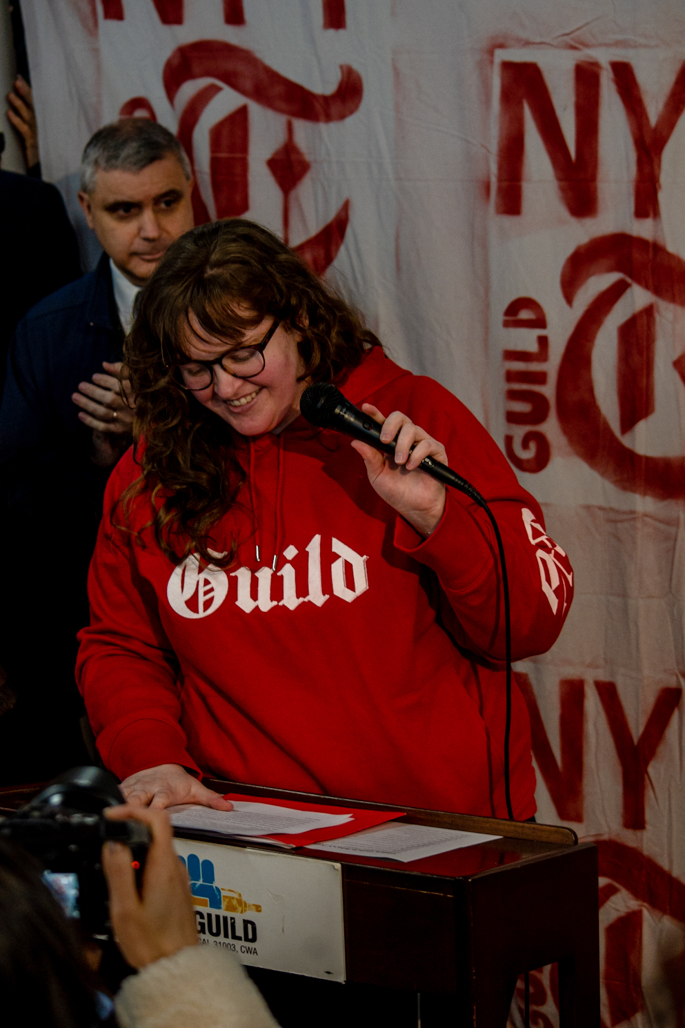 Carrie Price, a woman with short brown hair, standing at the podium wearing a red hoodie with “Guild” written on it.