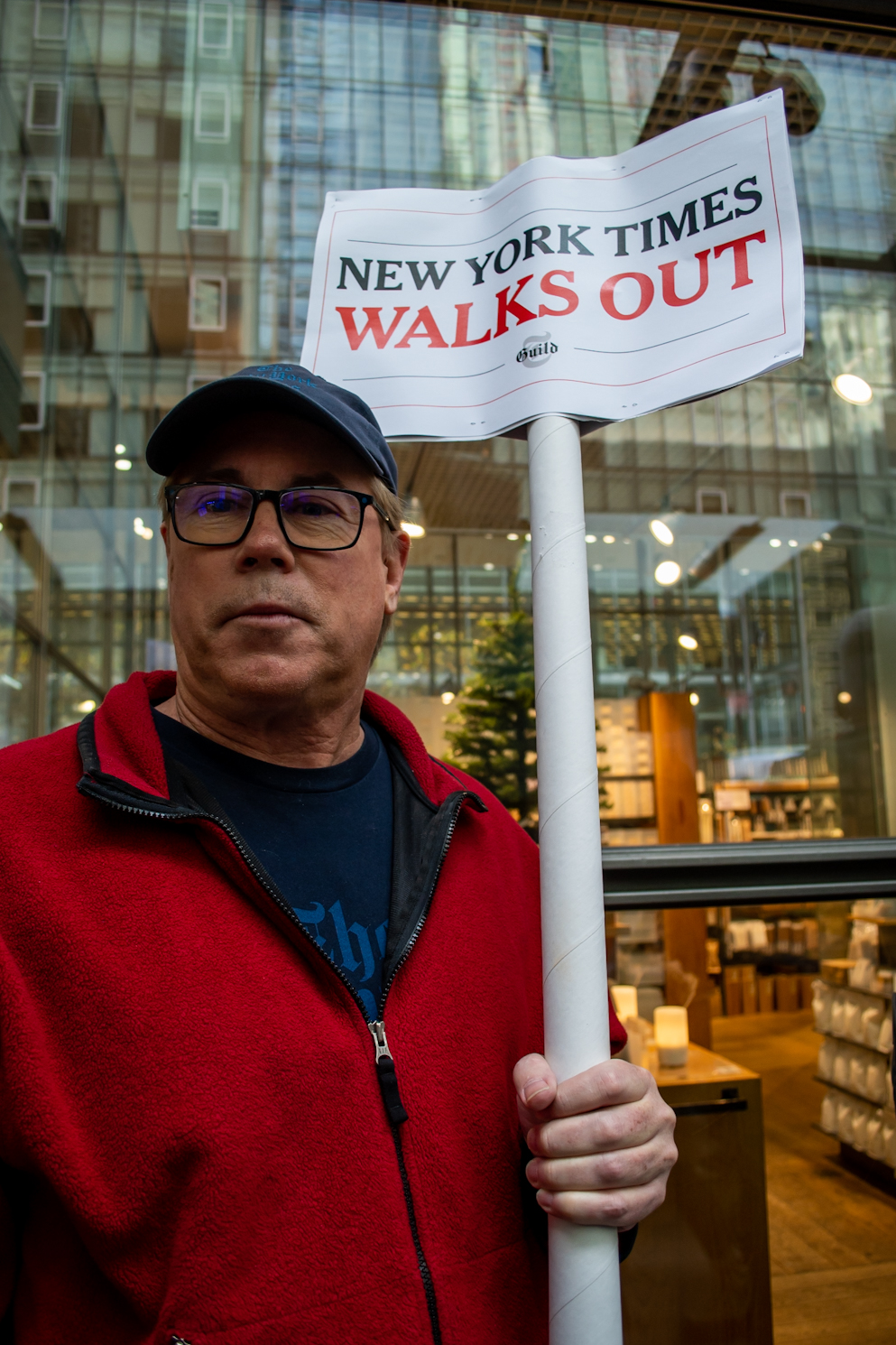 Patrick McGeehan, a man wearing a hat and a red jacket, holds a sign that reads “New York Times Walks Out.”