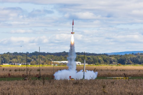 A red and white rocket launching off the ground in a wheat field.