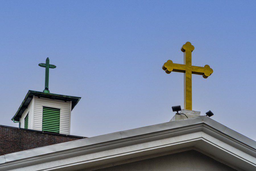 Green and gold crosses on the left and right respectively stand side by side on the roofs of mostly out-of-frame buildings against a clear, blue sky.