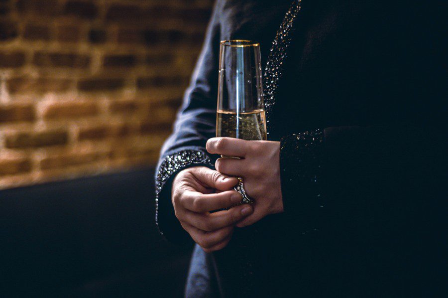 The torso of a person holding a champagne glass with a silver ring on their finger. The person wears a dark blue outfit with light blue gems.