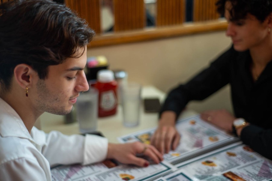 A dark-haired man with yellow highlights wearing a white shirt and a small, gold dangling earring with a red gem looks off into the distance while sitting to eat food at a diner with another dark-haired man.