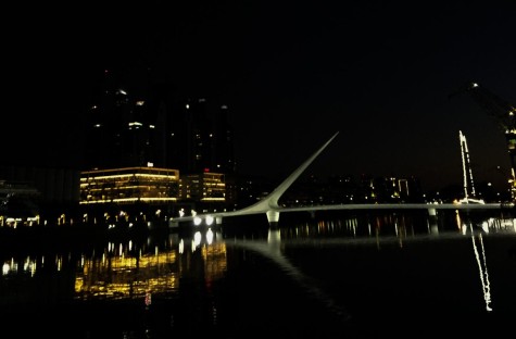 A bridge across a large body of water under the black night sky in front of a lit street around the water. There are facades of buildings visible only by the light coming from their windows.