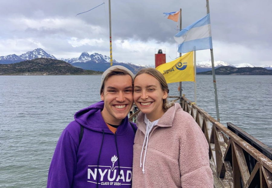 Two people smile while standing in front of a lake with snowy mountains on the horizon. On the left is a person wearing a purple hoodie with text “N.Y.U.A.D. Class of 2024.” On the right, the other person wears a pink jacket.