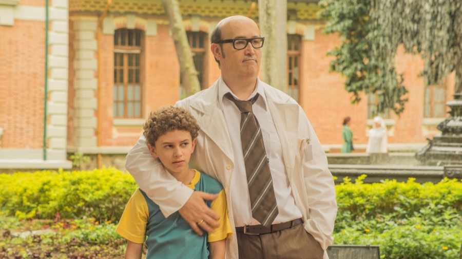 A father wearing glasses, a business formal outfit wraps his son, who is wearing a yellow and blue t-shirt, around his arm.