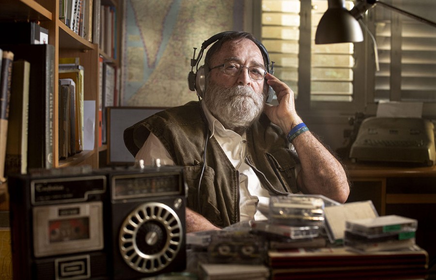 A man with thick, white facial hair sits behind a table filled with cassette tapes and a radio. He looks into the camera and wears a pair of glasses, a brown vest, a white shirt and a pair of headphones.