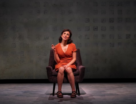 A woman wearing an orange dress sits in a sofa chair on stage in front of a gray backdrop with smudged square patterns on it.