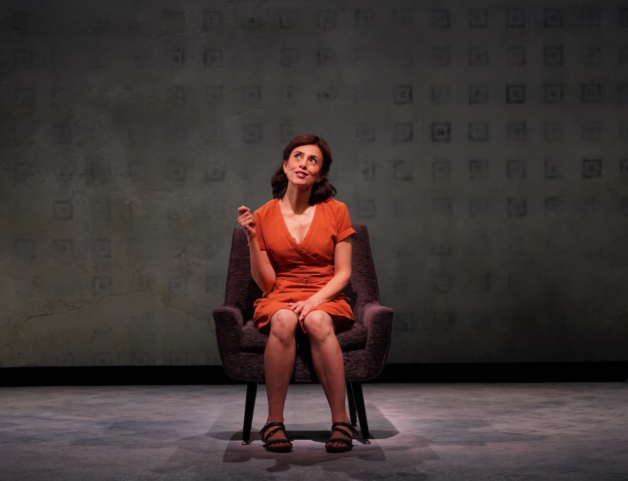 A+woman+wearing+an+orange+dress+sits+in+a+sofa+chair+on+stage+in+front+of+a+gray+backdrop+with+smudged+square+patterns+on+it.