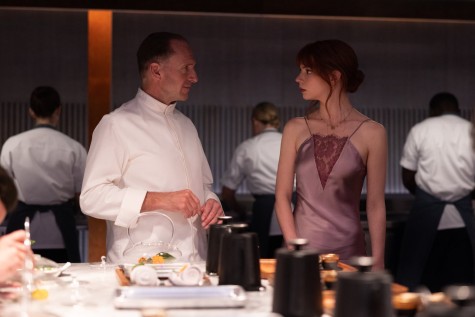 A male wearing a white chef uniform and a female wearing a purple silk dress look at each other. They are standing in an open kitchen with several cooks working in the back and many containers for food put on a table in front of them.