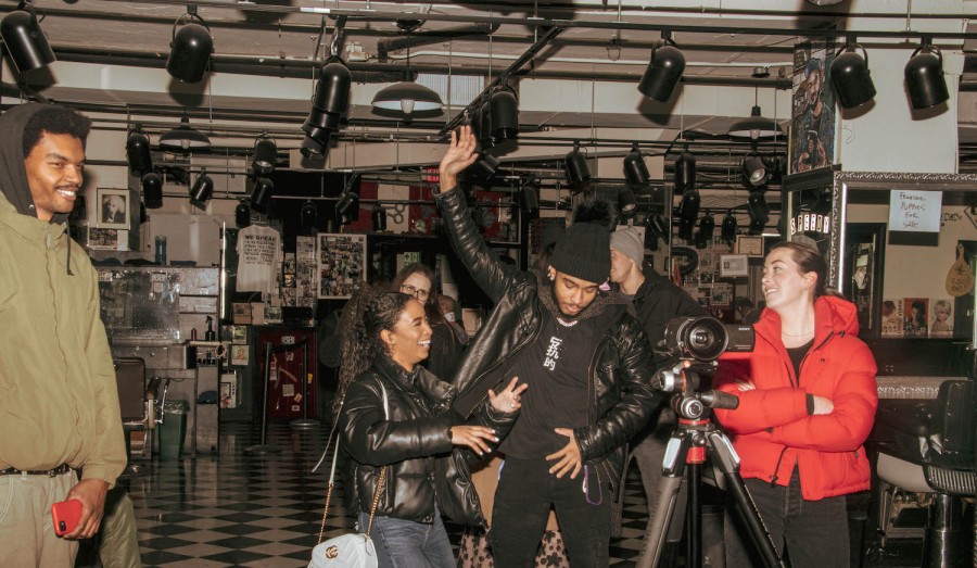 From left to right: a man wearing a green hoodie, a woman wearing a black puffer jacket, a man wearing a black leather jacket and a woman wearing a red puffer jacket dancing in a room. There is a tripod with a camera in front of them.