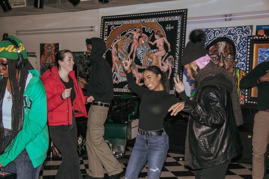 From left to right: a woman wearing a green hoodie, a woman wearing a black puffer jacket, a man wearing a black leather jacket and a woman wearing a red puffer jacket dancing in a room. There is artwork on the wall behind them.