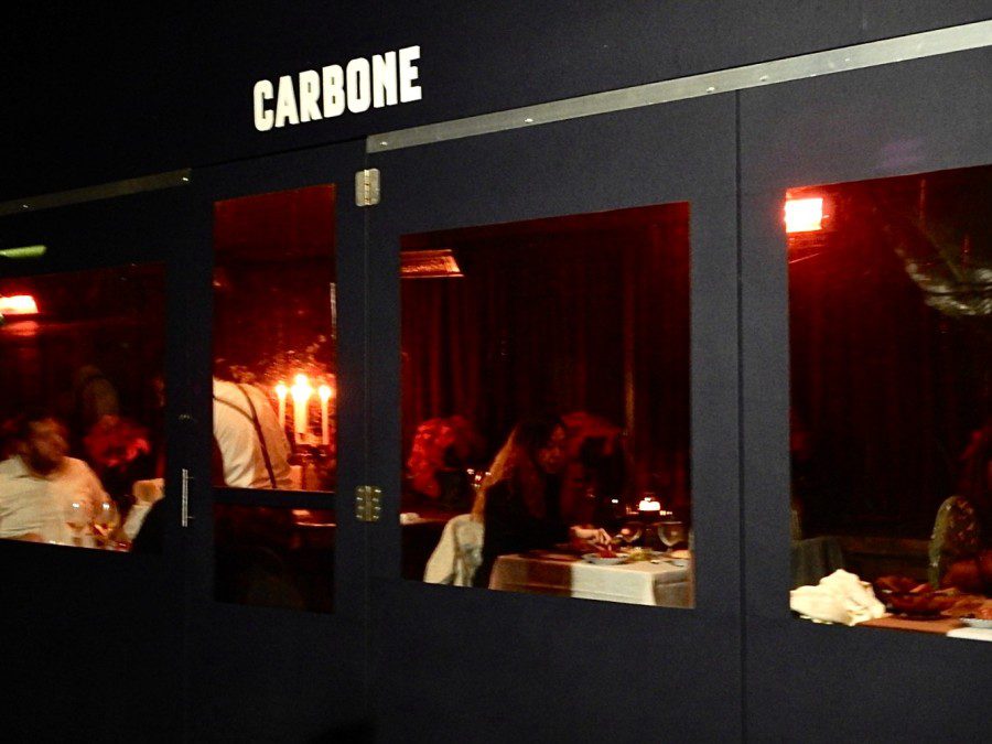 A black facade that reads “Carbone” in white font. Inside, there is dim red lighting. A female is visible sitting down alone at a table in the restaurant.