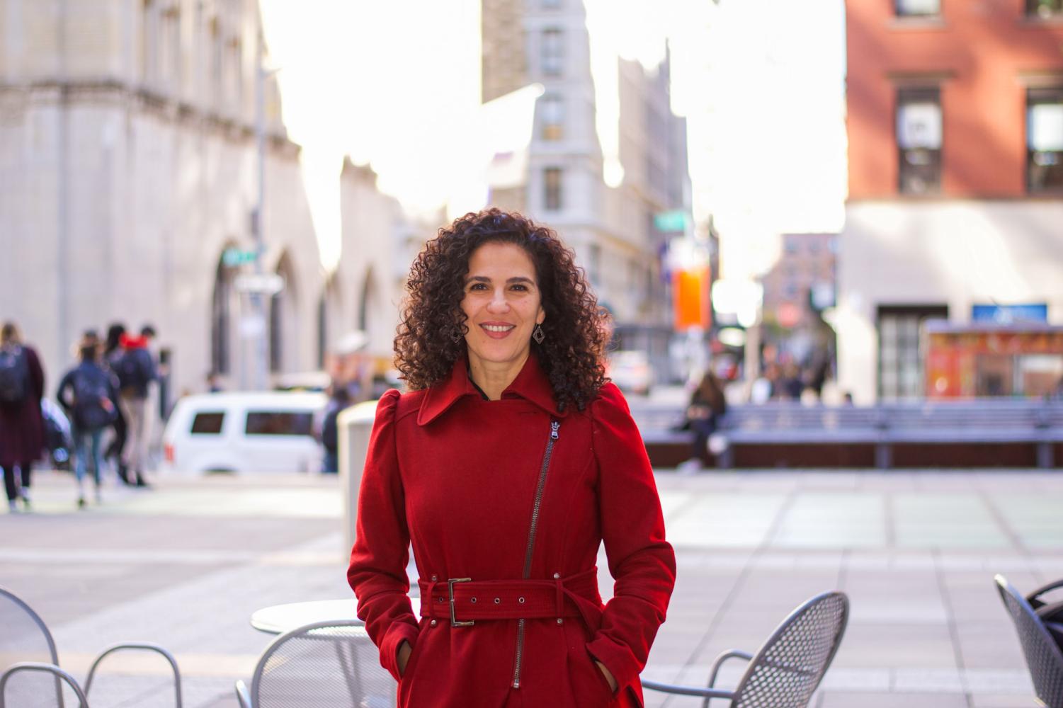 A female with black curly hair wears a long red coat and is smiling and looking toward the camera.