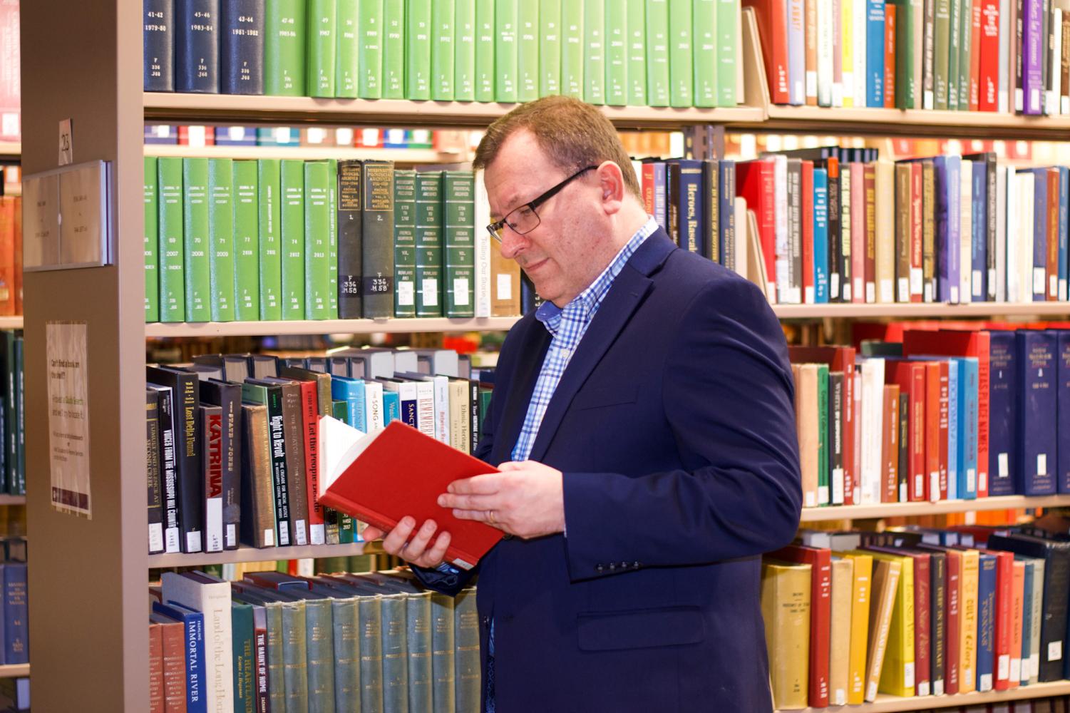 A male wearing glasses, a navy blue blazer and a blue shirt holds a red book with his hands. Behind him is a bookshelf with multiple rows of books with red, green, blue, black and brown covers.