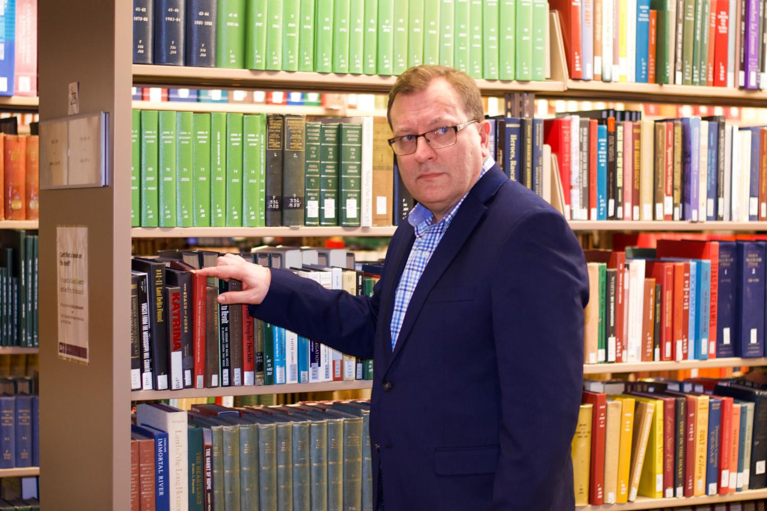 A male wearing glasses, a navy blue blazer and a blue shirt holds the tip of a red book with his right hand. This book is placed on a bookshelf that has multiple rows of books with red, green, blue, black and brown covers.