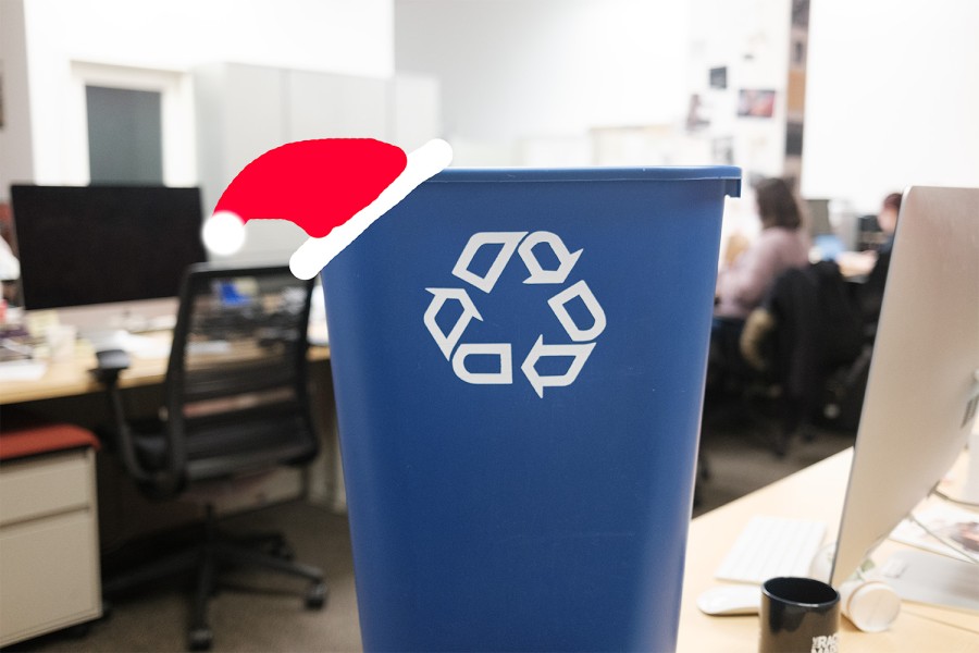 A blue, plastic recycling bin with a white U+2672 Universal Recycling Symbol printed on it. A red Santa hat illustration is overlaid on the recycling bin.