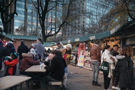 A series of green-roofed booths selling various foods are visited by people wearing brown, black and white coats. They are buying products and sitting at beige picnic tables drinking out of white cups.