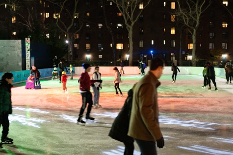 Sixteen people in various colored coats skating across a heavily scratched ice skating rink at night. The surface is covered in orange and green light. Four children wearing brightly colored puffy coats are being led across the ice by parents, one with a purple ice skating assistance device.