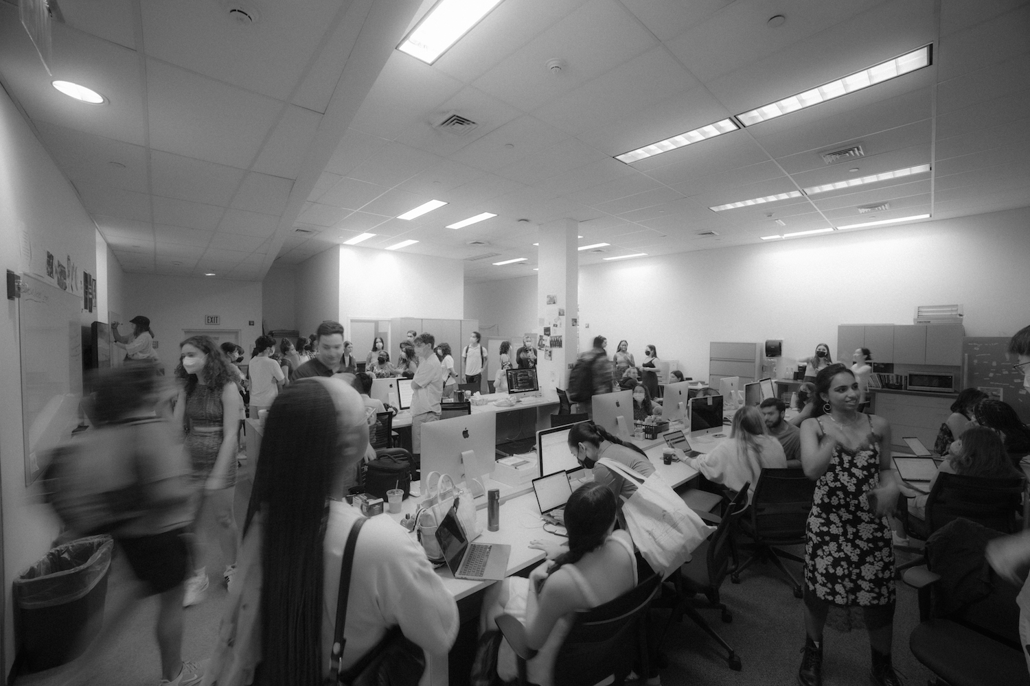 An office space with no windows and white walls. People sit at desks and walk around.