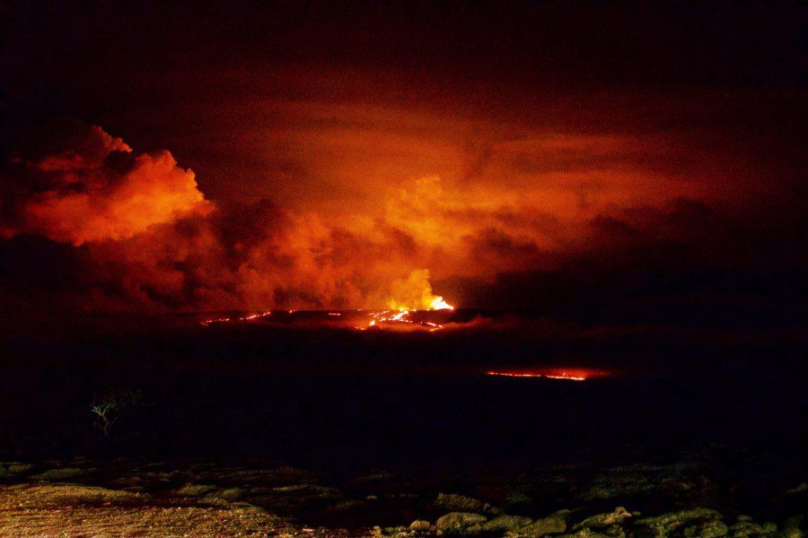The Mauna Loa erupts against a dark sky. The lava in bright red and orange lights up the volcano and tints the color of smoke shooting out from the crater.