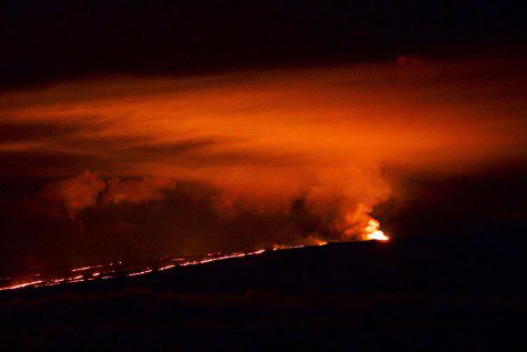 The Mauna Loa erupts against a dark sky. The lava in bright red and orange lights up the volcano and tints the color of smoke shooting out from the crater.