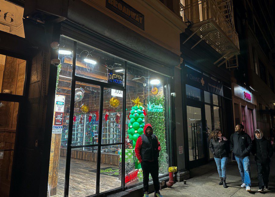 A storefront, viewed from the street outside, with harsh white lighting, and rows of colorful cannabis products displayed inside. A man wearing a red hoodie and black vest stands outside.