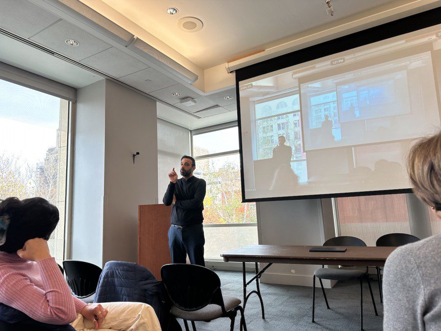 Professor Rami Salameh stands in front of a podium, speaking to an audience in the Kimmel Center for University Life, while a projector screen behind him displays a Zoom virtual meeting.