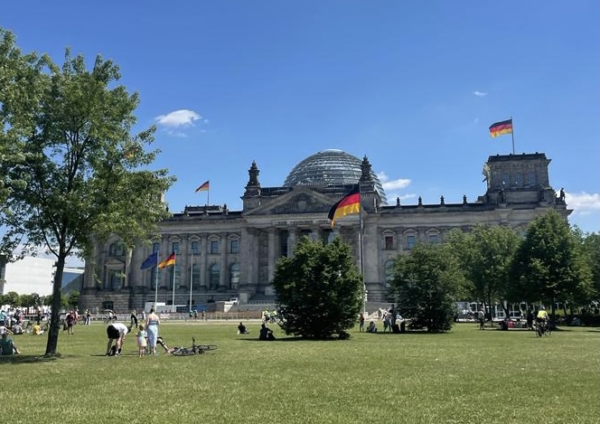 A large gray building with a complex dome in its center sitting in front of a lush park with multiple trees and people.