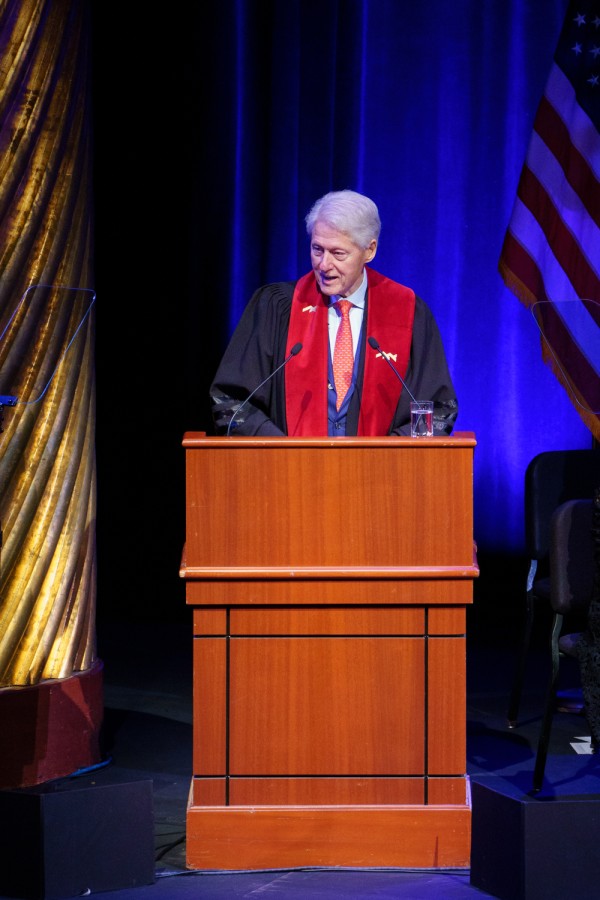 Bill Clinton wears a red tie with a blue robe as he speaks behind a wooden podium on a stage with a blue backdrop behind him.
