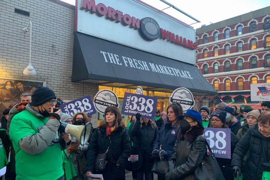 A group of protestors wearing green T-shirts rally in front of s Morton Williams supermarket. They are holding signs that read “LOCAL 338 R.W.D.S.U/U.F.C.W.”