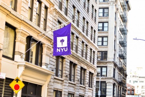 A photo of a purple flag that hangs from a flagpole attached to a brown building. The flag reads “NYU” in white-colored font.