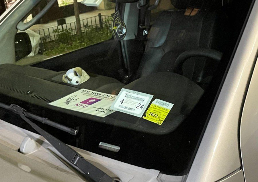 A gray car with a yellow sticker and a white sticker in the bottom right corner of its windshield. The stickers read 4 and 24 respectively. The car also has a placard that reads New York University displayed.